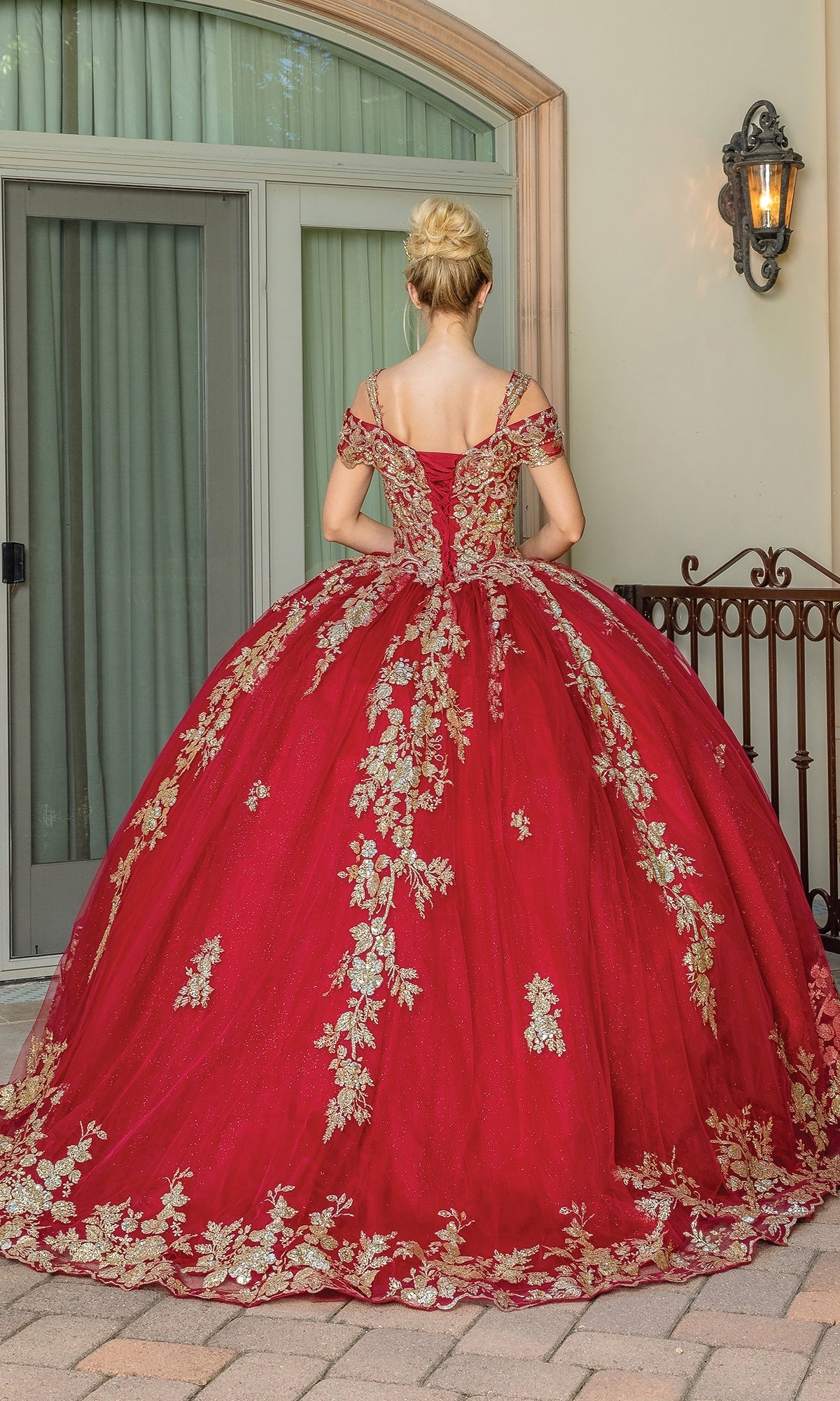 A woman wearing the Burgundy Red Quinceanera Dress 1657, featuring gold lace embroidery, stands with her back to glass doors and a wall-mounted lantern.